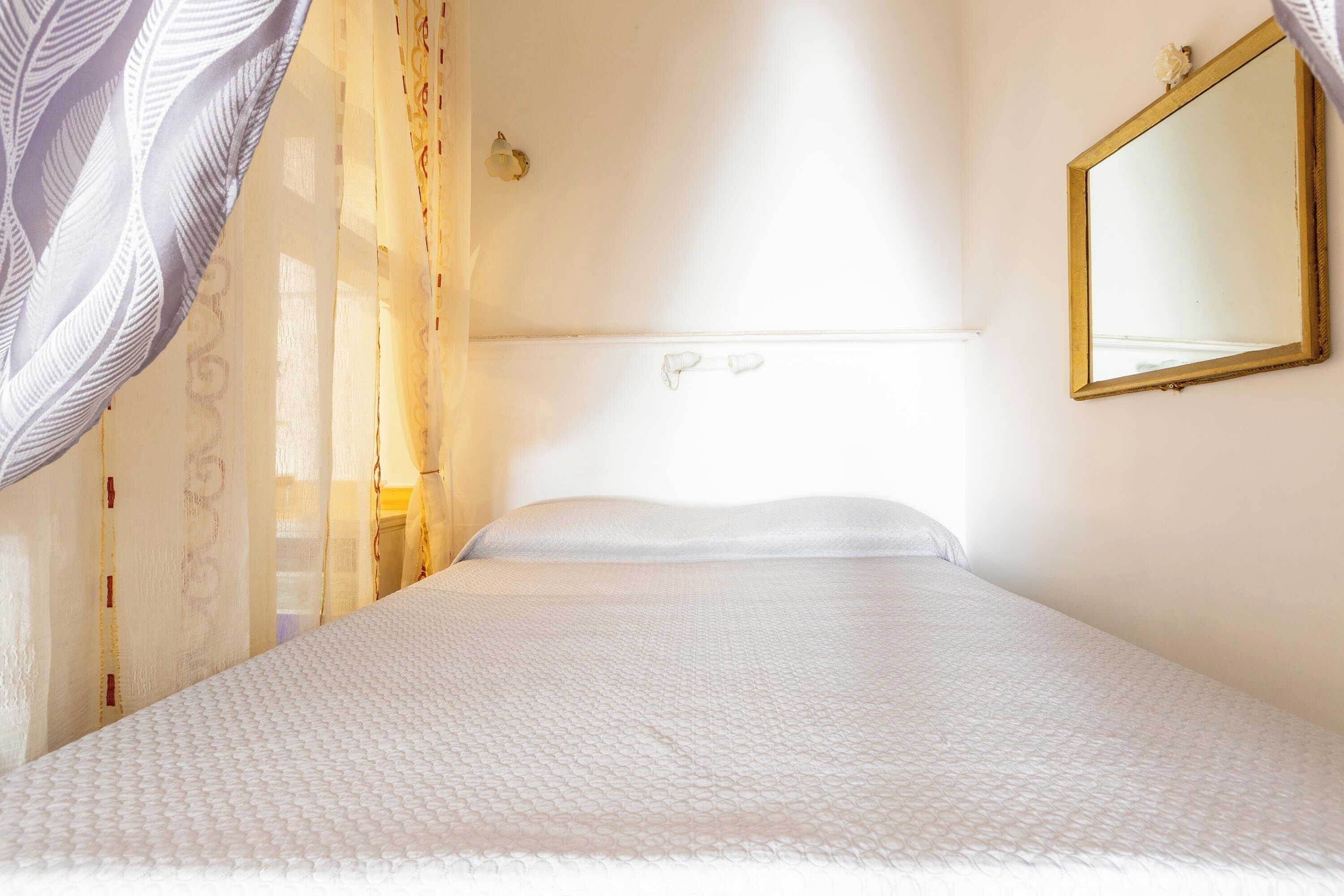 A neatly made bed with a white bedspread and pillows, a wall-mounted mirror, and a window with sheer curtains.