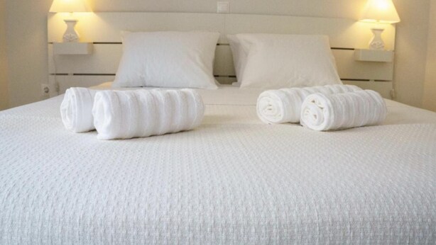 Una cama bien hecha con sábanas y almohadas blancas, dos toallas enrolladas y dos lámparas de noche sobre mesitas blancas.
