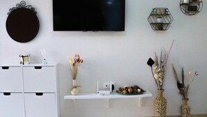 Una sala de estar con una televisión de pantalla plana montada en la pared, un mueble blanco con tiradores negros, un estante blanco con adornos y dos jarrones grandes con plantas secas.