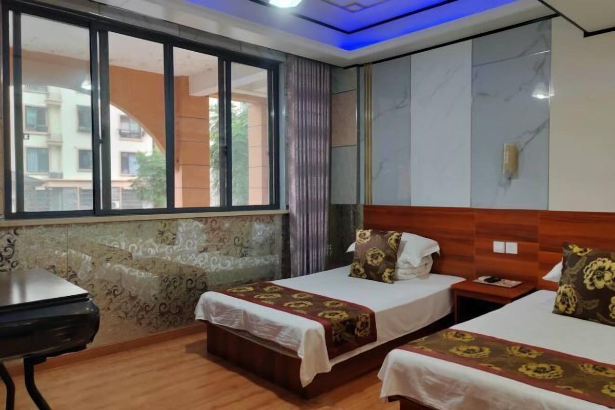 A hotel room with two beds, a small table, a window with a view of buildings, and a patterned wall.