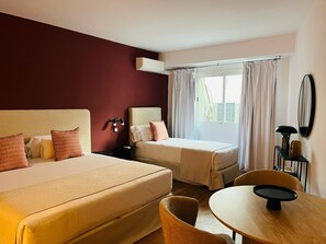 Habitación de hotel con dos camas, una mesita, una silla, un escritorio con lámpara y una ventana con cortinas.
