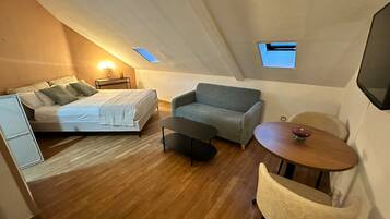 Una habitación compacta y moderna con una cama, un sofá gris, una pequeña mesa redonda y una silla.