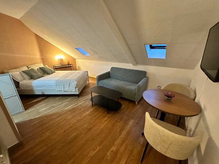 Una habitación compacta y moderna con una cama, un sofá gris, una pequeña mesa redonda y una silla.