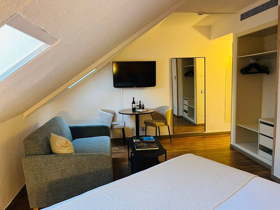 A modern room with a sloped ceiling, a flat-screen TV mounted on the wall, a grey sofa with a white cushion, a small round table with a bottle and a book, and a walk-in closet with a mirror and hanging clothes.