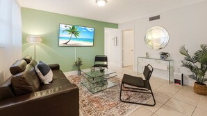 A living room with a leather sofa, a glass coffee table, a TV displaying a beach scene, a round mirror, and a potted plant. - Comfortable 3-bedroom apartment with easy access to major highways (MIAMI)