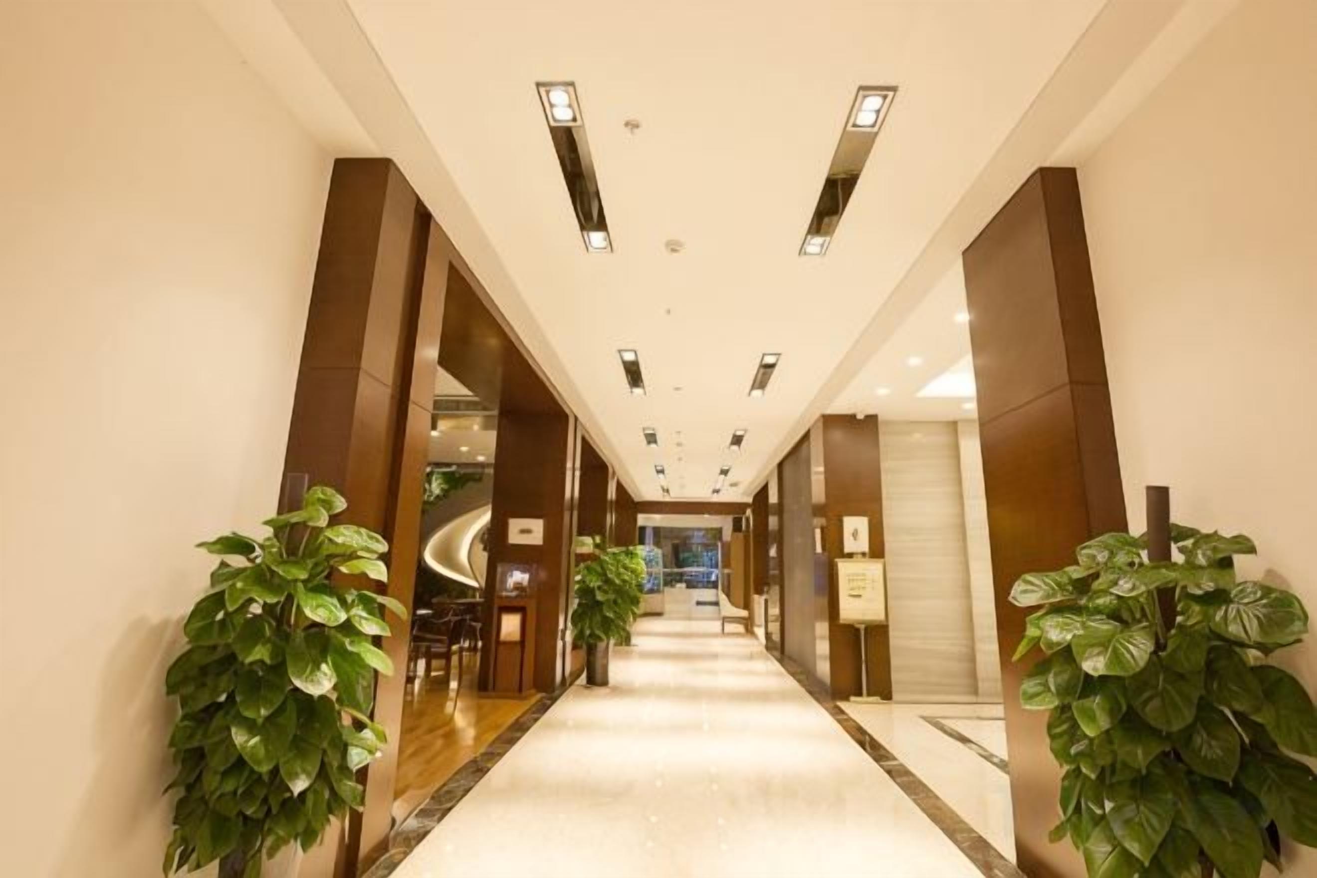 Un pasillo de hotel con piso de mármol, plantas en macetas grandes y un diseño moderno de techo.