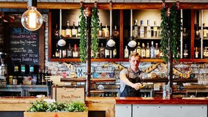Un bar avec un comptoir en bois, des étagères remplies de bouteilles d’alcool et un tableau noir affichant la carte des boissons.