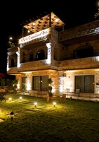 Un hotel su due piani con facciata in pietra, un livello superiore coperto e una zona esterna ben illuminata con posti a sedere.