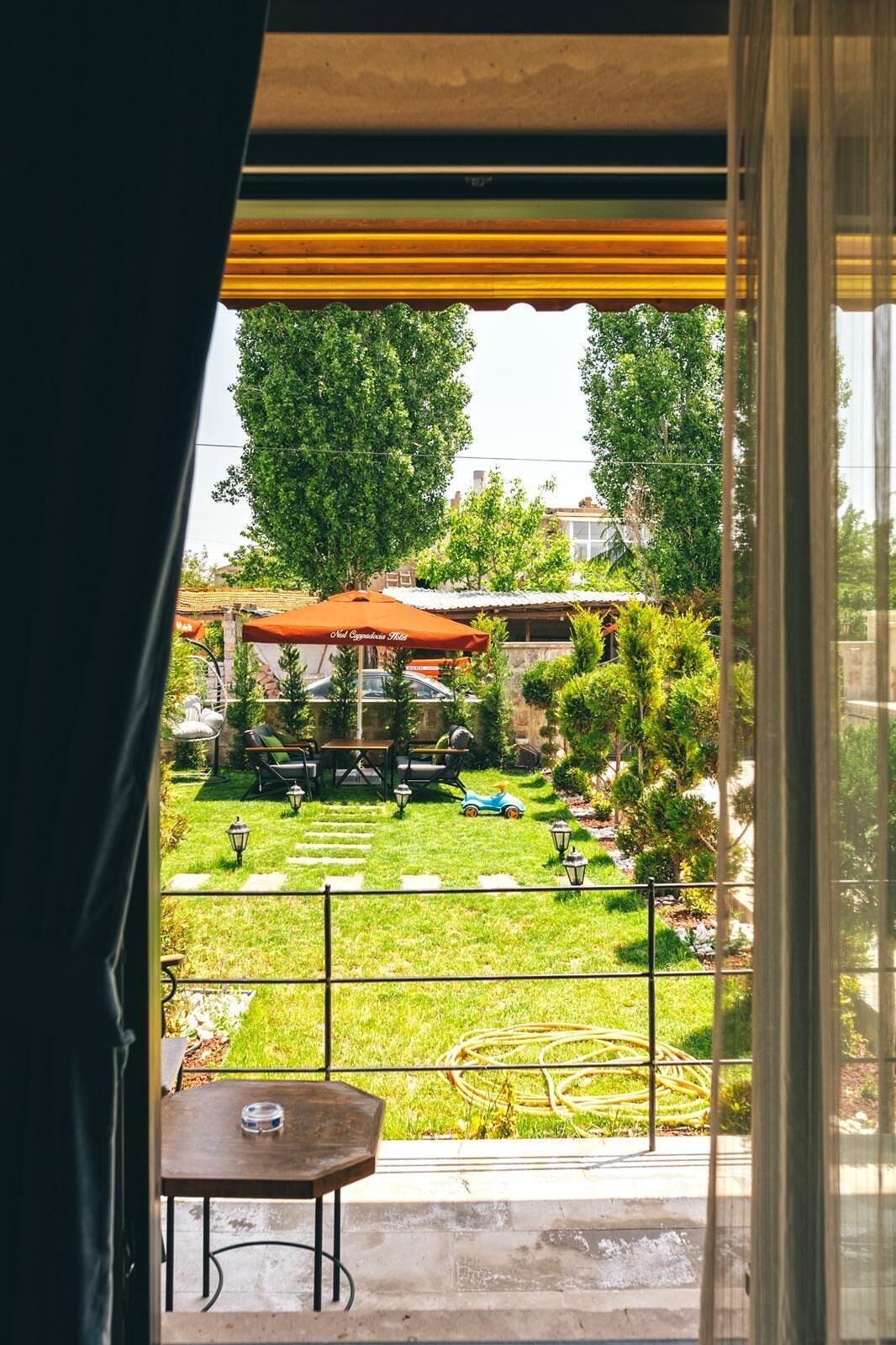 A view from a balcony with a table and chairs, a small garden with a lawn, a play area with a toy car, and a building with a red awning labeled "Hotel Cagaba Hotel".