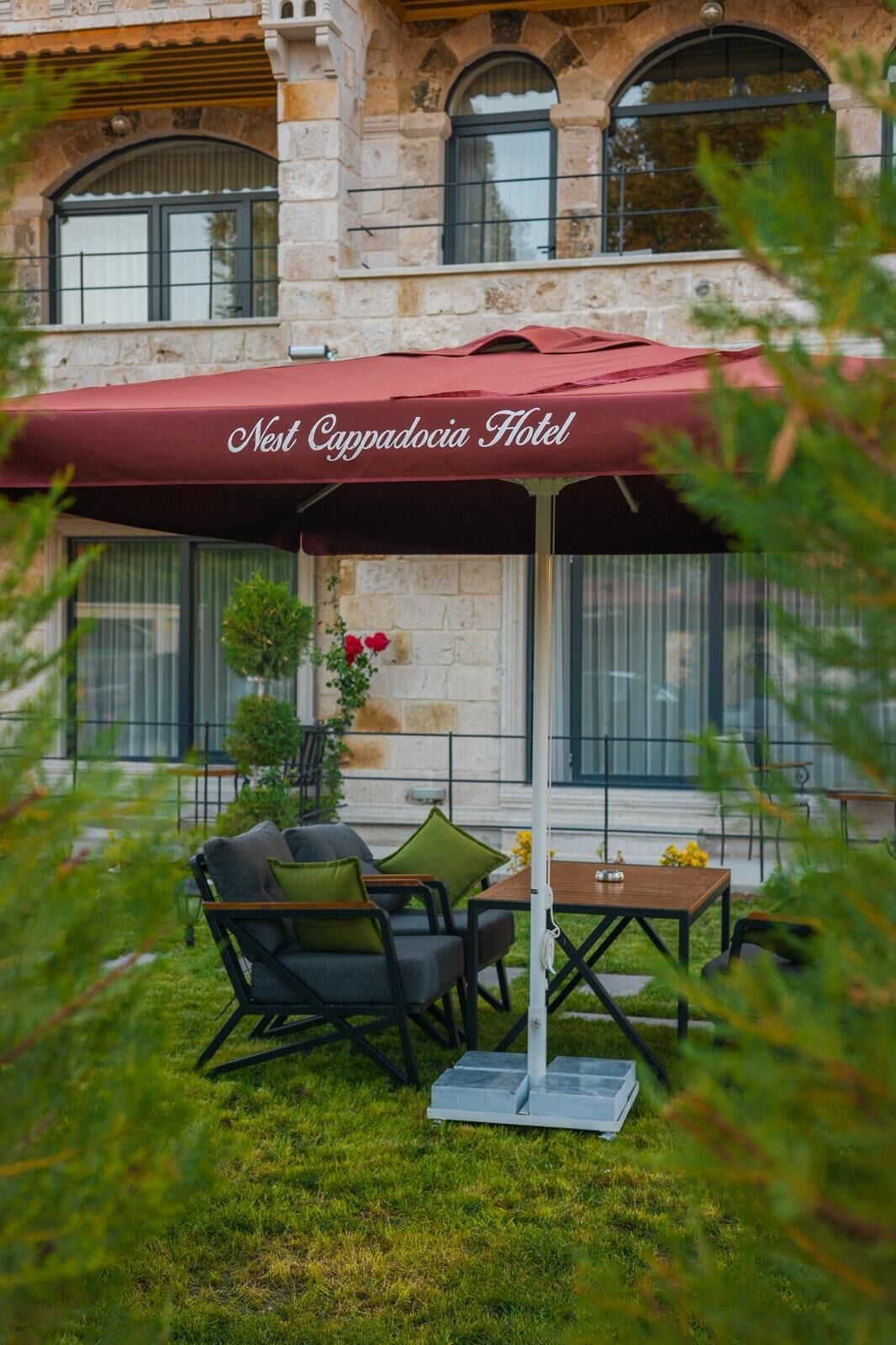Een terras met een rode parasol, gelabeld 'Nest Cappadocia Hotel', met een tafel, stoelen, een kleine potplant en een stenen gebouw op de achtergrond.
