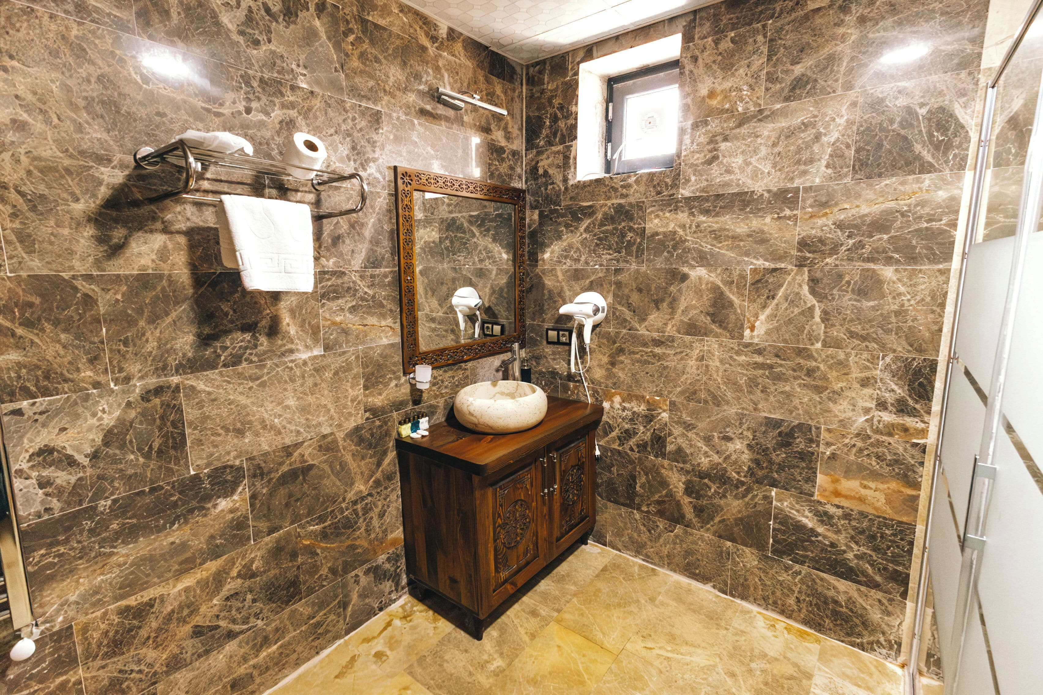 Un baño con pared de mármol, tocador de madera, un espejo grande y una ventana.