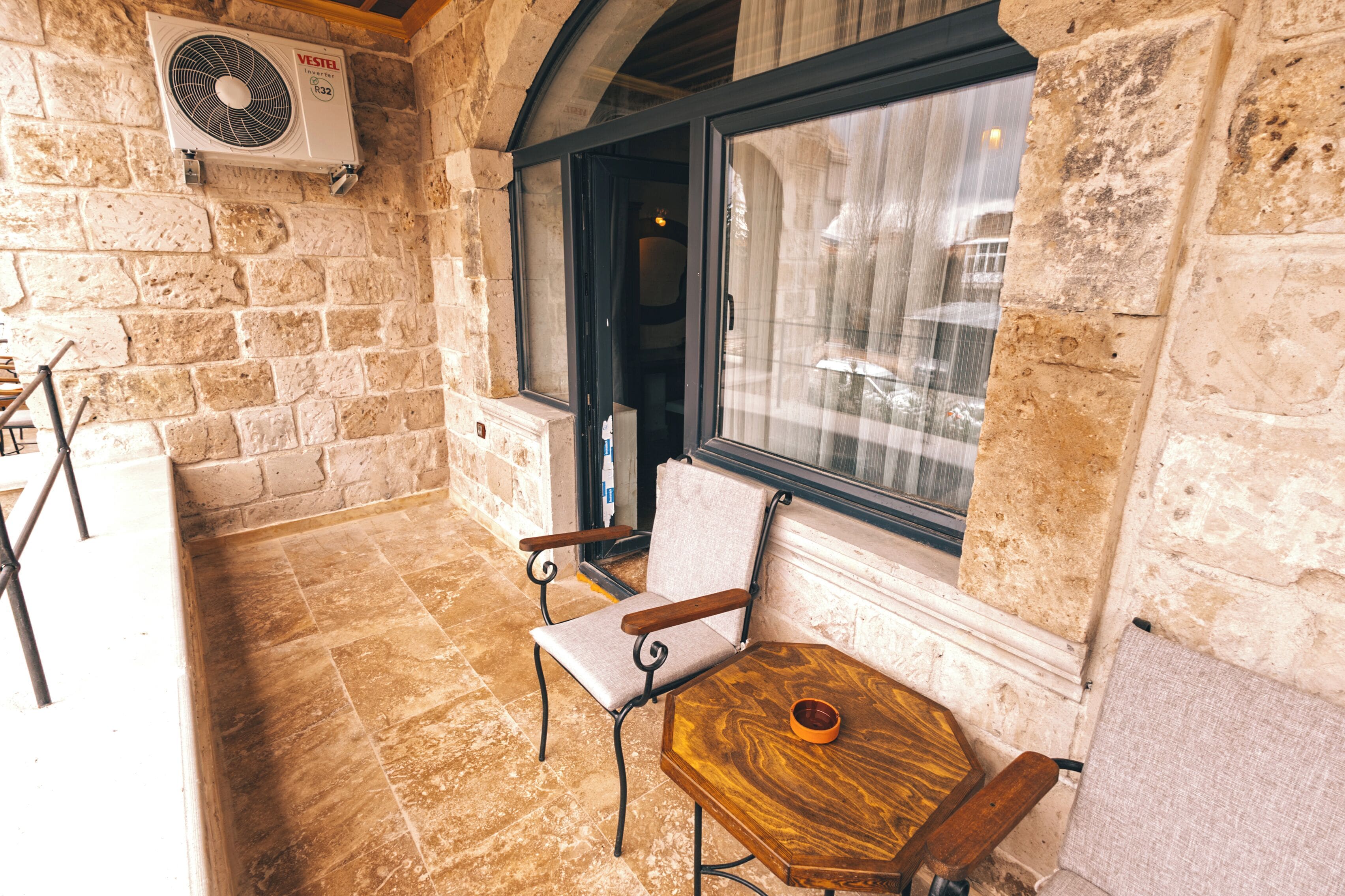 A stone-walled room with a window, a fan, a wooden table, and two chairs.