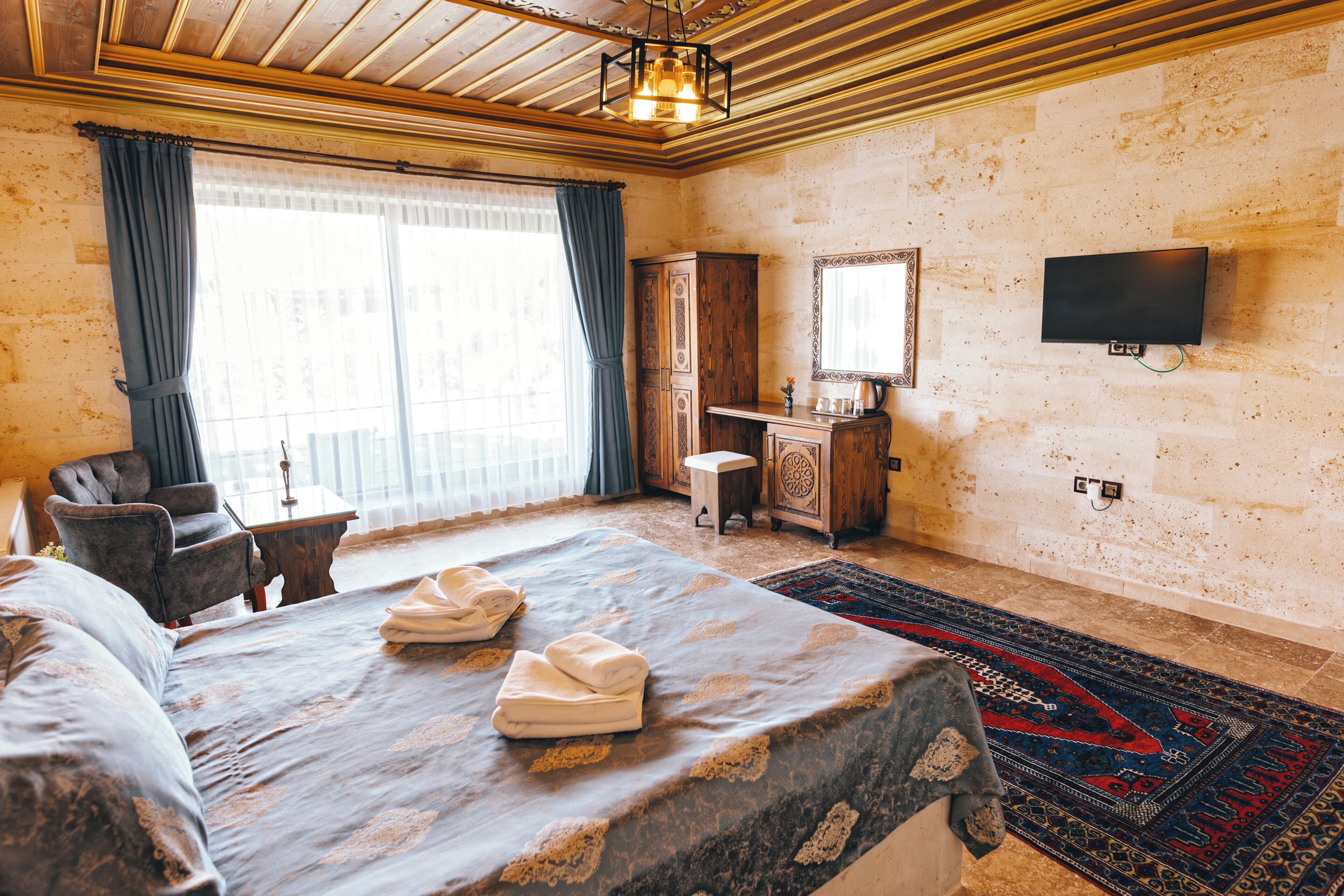 A hotel room with a bed, a desk, a chair, a wardrobe, a television, and a large window with curtains.