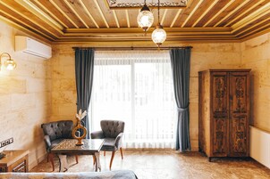 Una habitación con techo de madera, un ventanal con cortinas, dos sillones, una mesa con un florero y un mueble de madera.