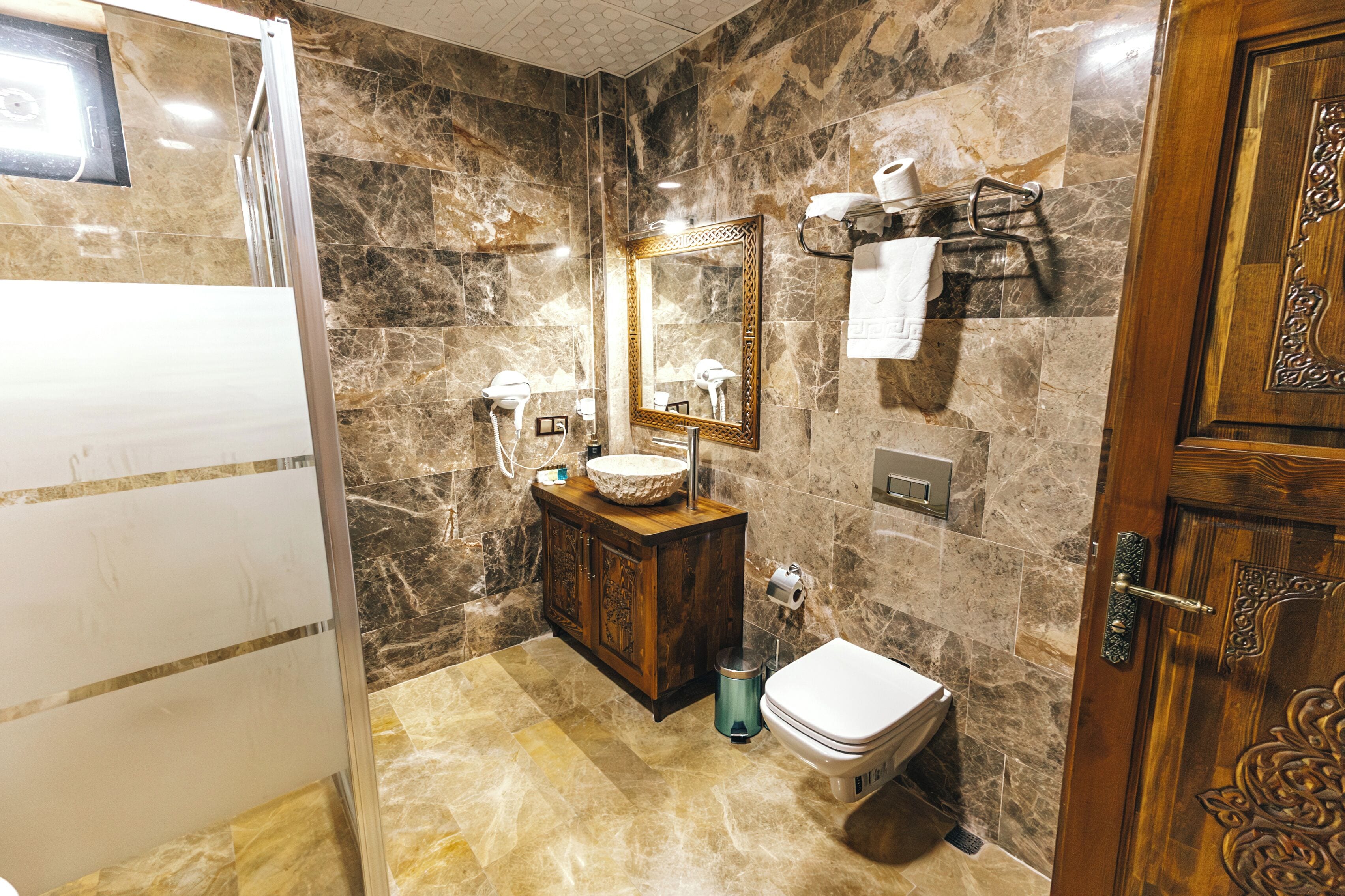 A bathroom with marble walls, a wooden vanity, a sink, a toilet, a mirror, and a towel rack.