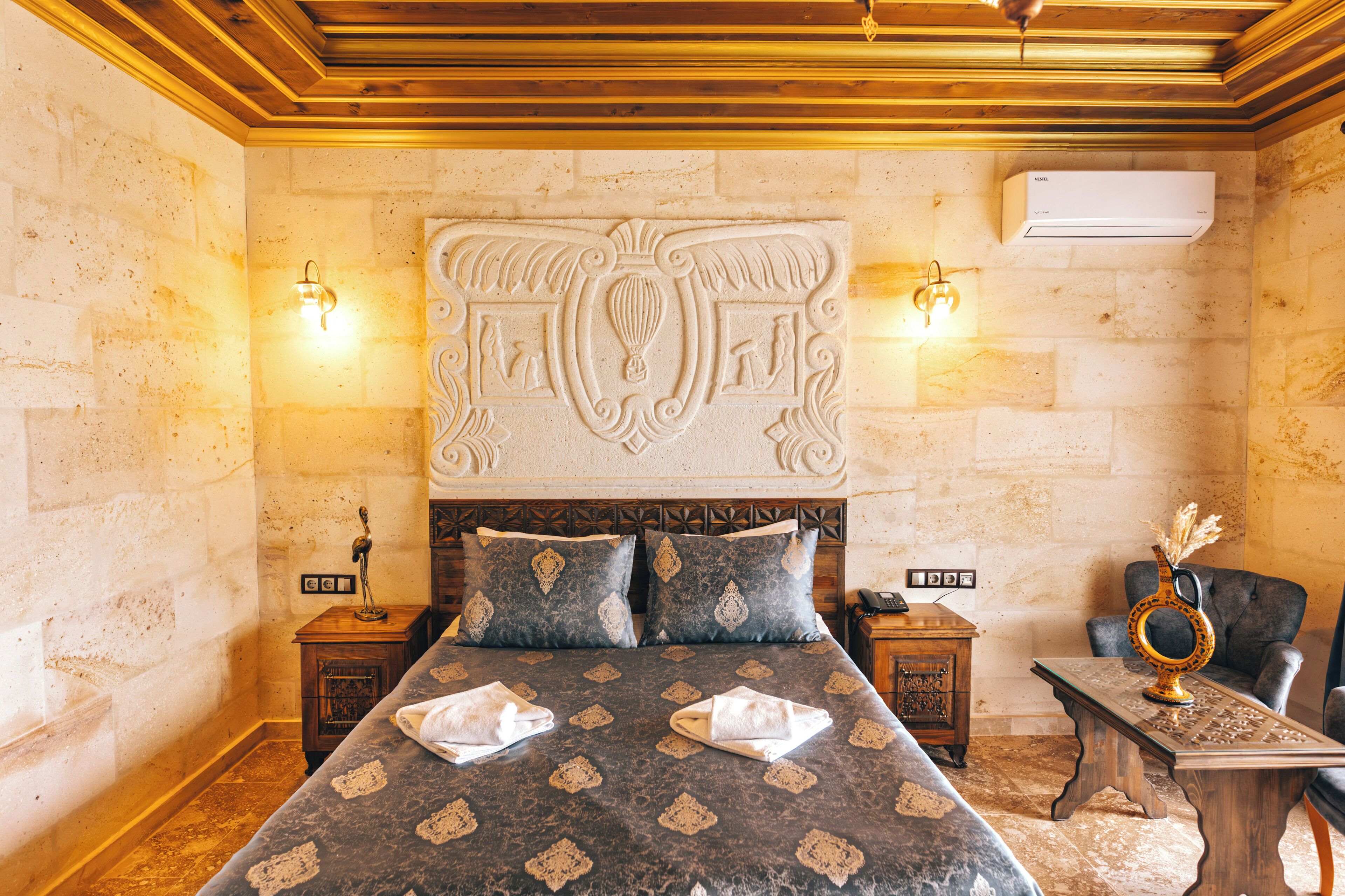 Quarto de hotel com uma cama grande, duas mesas de cabeceira, um painel de madeira na cabeceira e uma pequena mesa com um vaso.