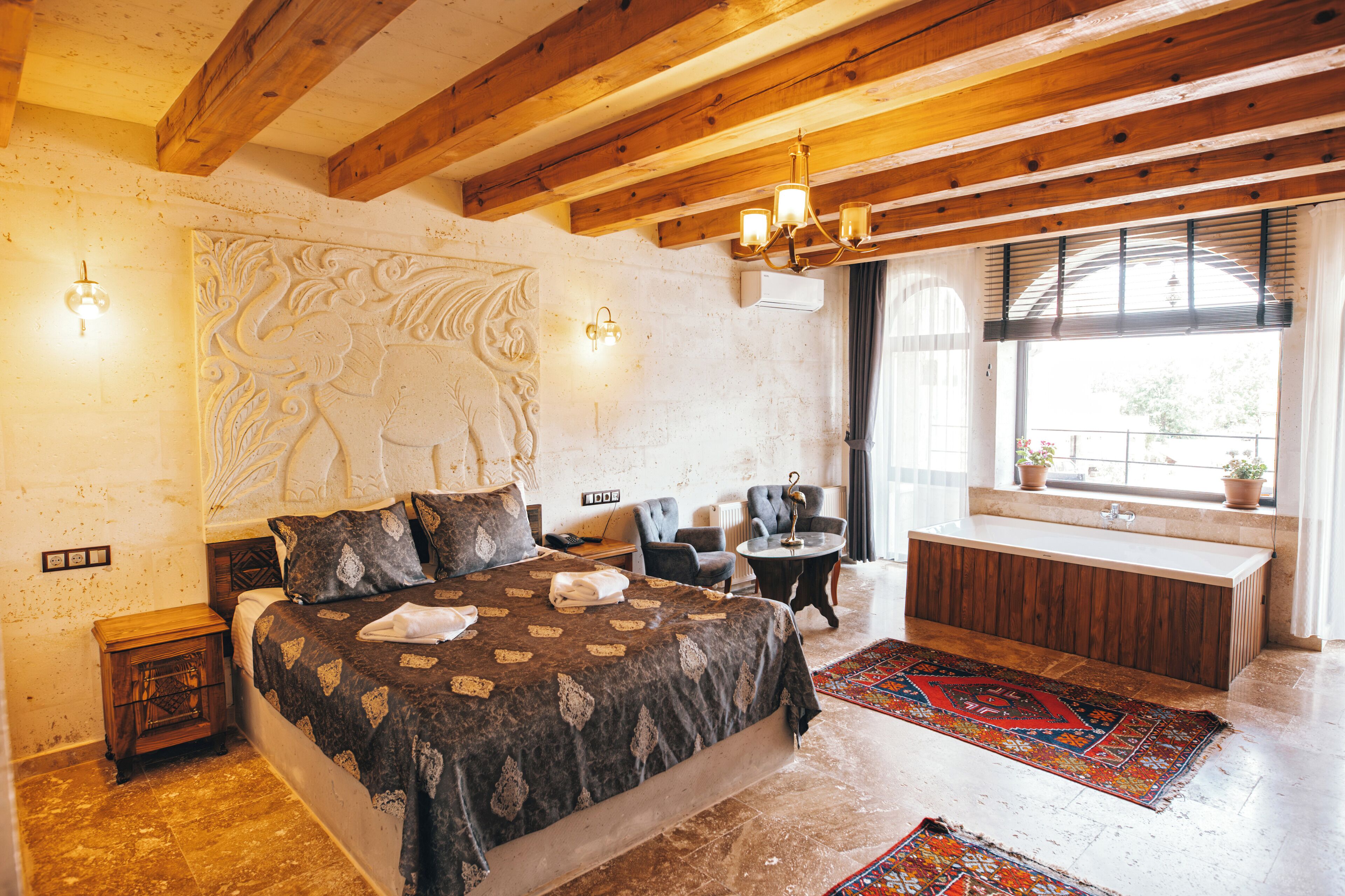 A bedroom with a large bed, a wooden ceiling, a small round table, a chair, and a window with a view.