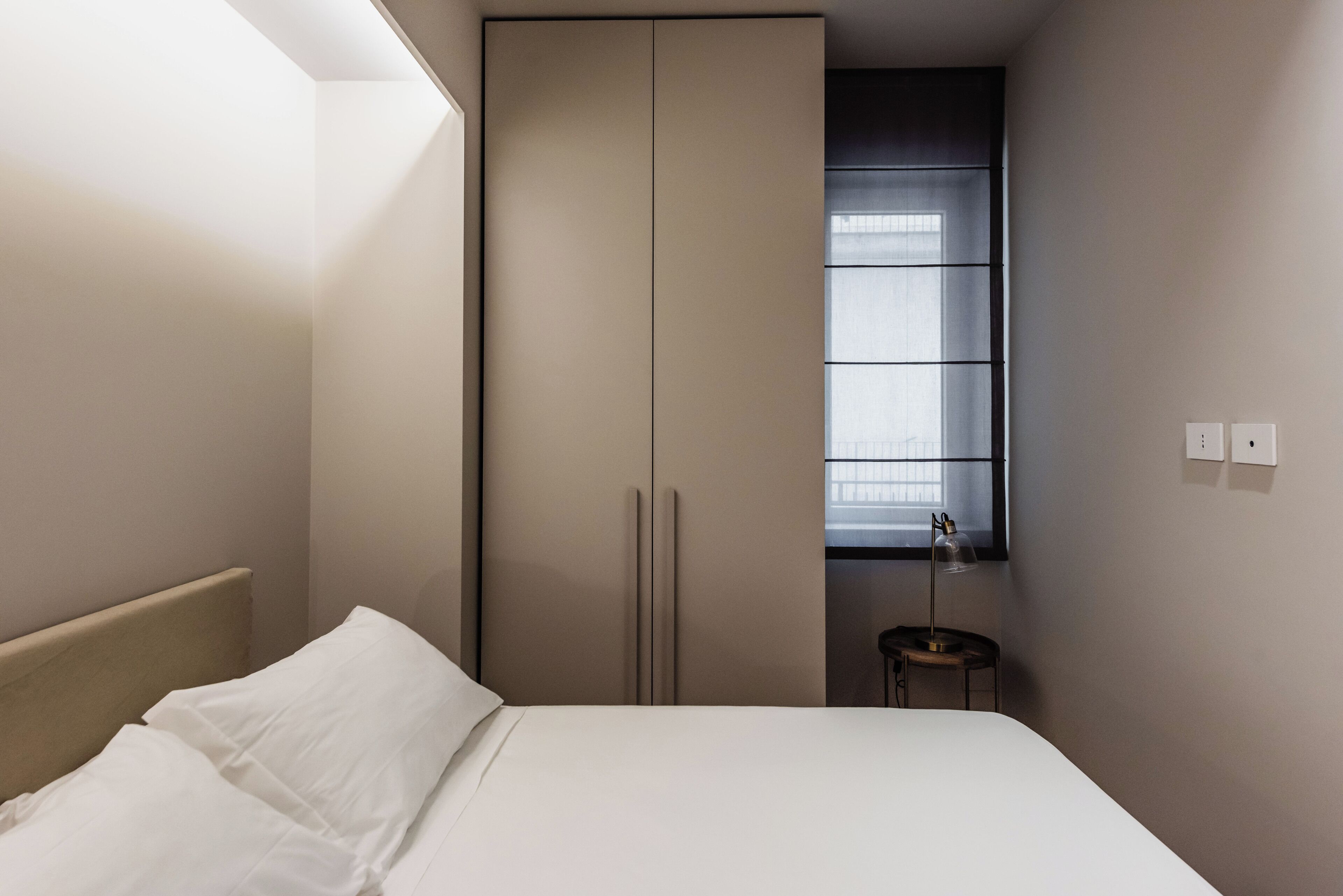 A bedroom with a bed, a wardrobe, a small table, and a window.