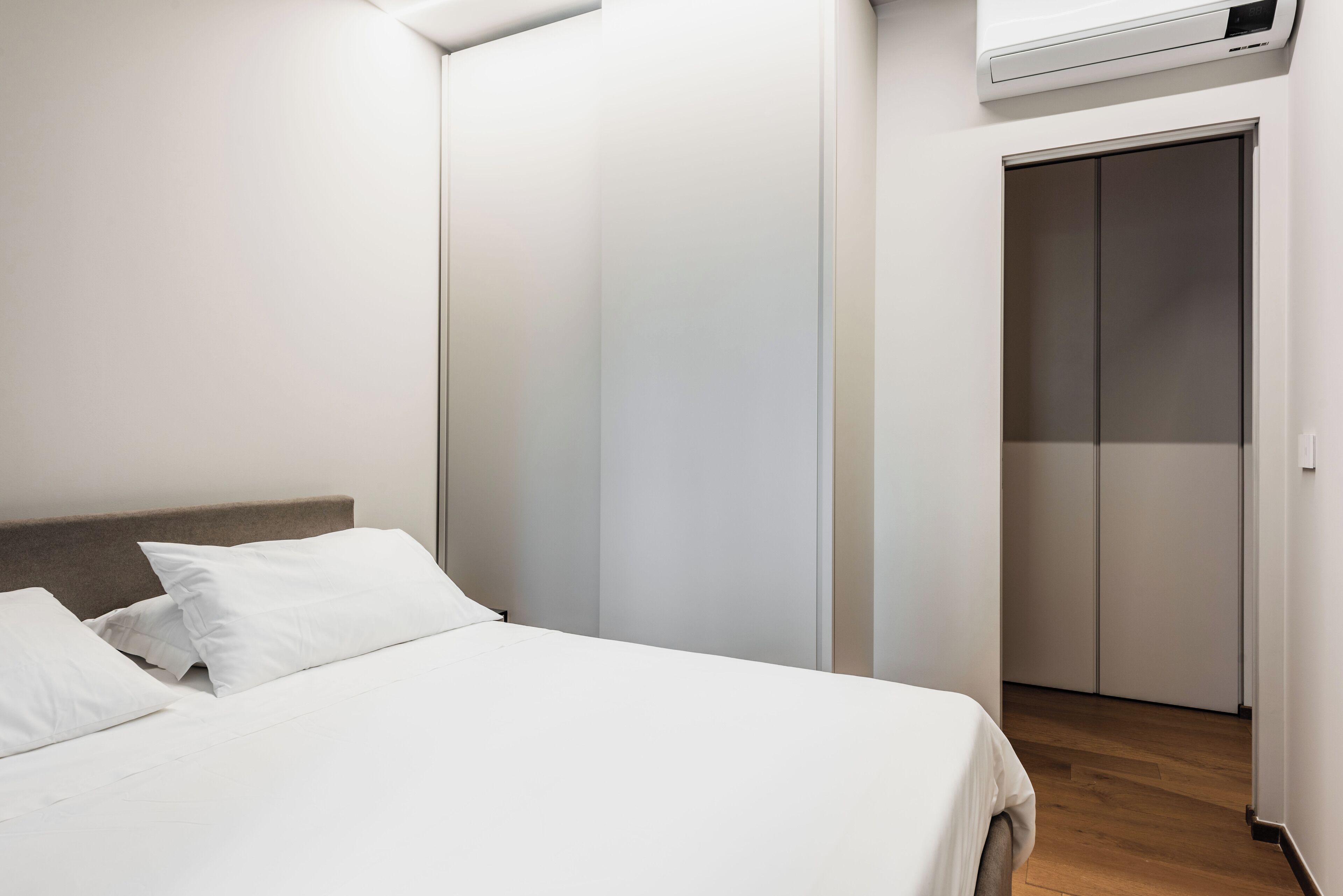 A bedroom with a bed, white bedding, a wooden headboard, a wardrobe, and a wall-mounted air conditioner.