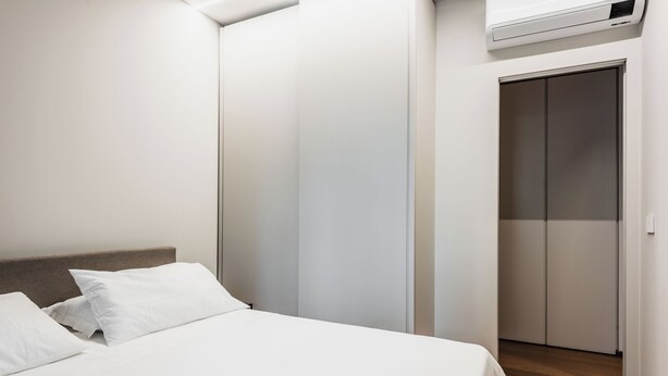 A bedroom with a bed, white bedding, a wooden headboard, a wardrobe, and a wall-mounted air conditioner. - Ferrini Home - Riso 74 (Catania)