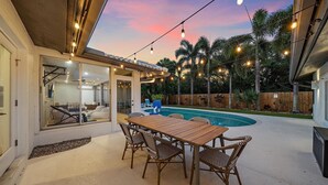 An outdoor patio with a wooden table and wicker chairs, a swimming pool, and string lights.