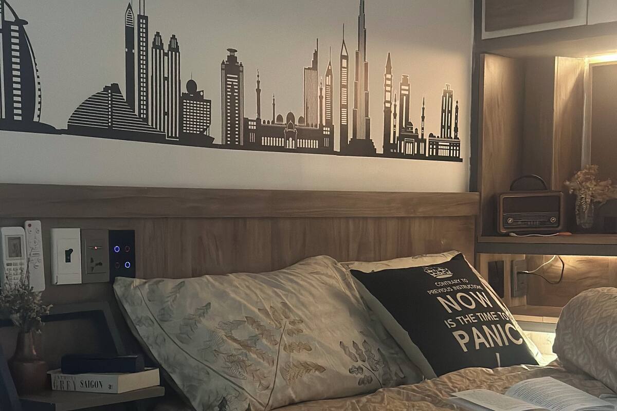 Un dormitorio con un mural de un paisaje urbano, una cama con ropa de cama estampada, una mesita de noche con libros y una lámpara, y una mesita de noche con un jarrón y un control remoto.