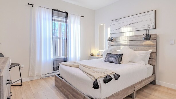 Un dormitorio con cama de madera, una mesita de noche, una ventana con cortinas translúcidas y una obra de arte en la pared.