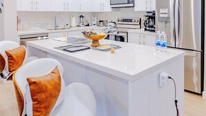 Una cocina moderna con armarios blancos, electrodomésticos de acero inoxidable, una isla central y una zona de estar con sillas blancas y cojines anaranjados.