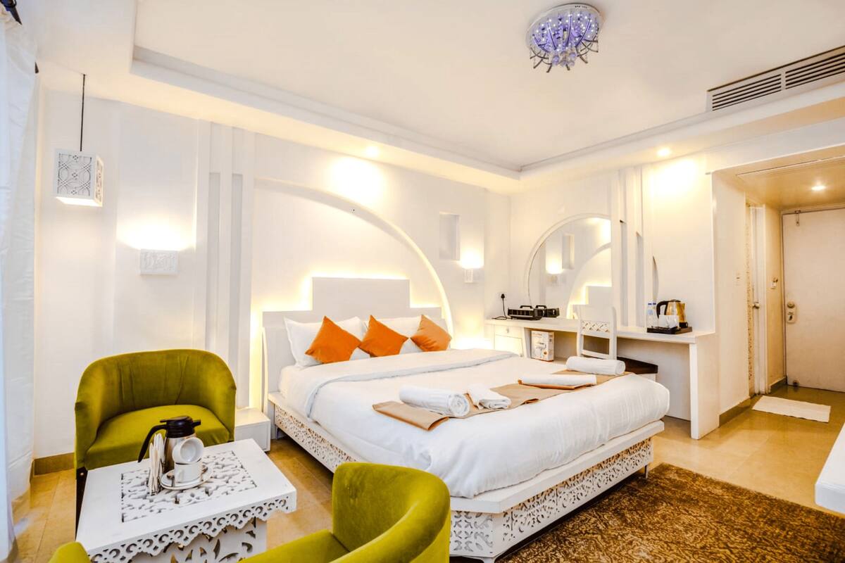 Une chambre d’hôtel avec un grand lit, deux fauteuils verts, une petite table avec une machine à café et un miroir au-dessus du lit.