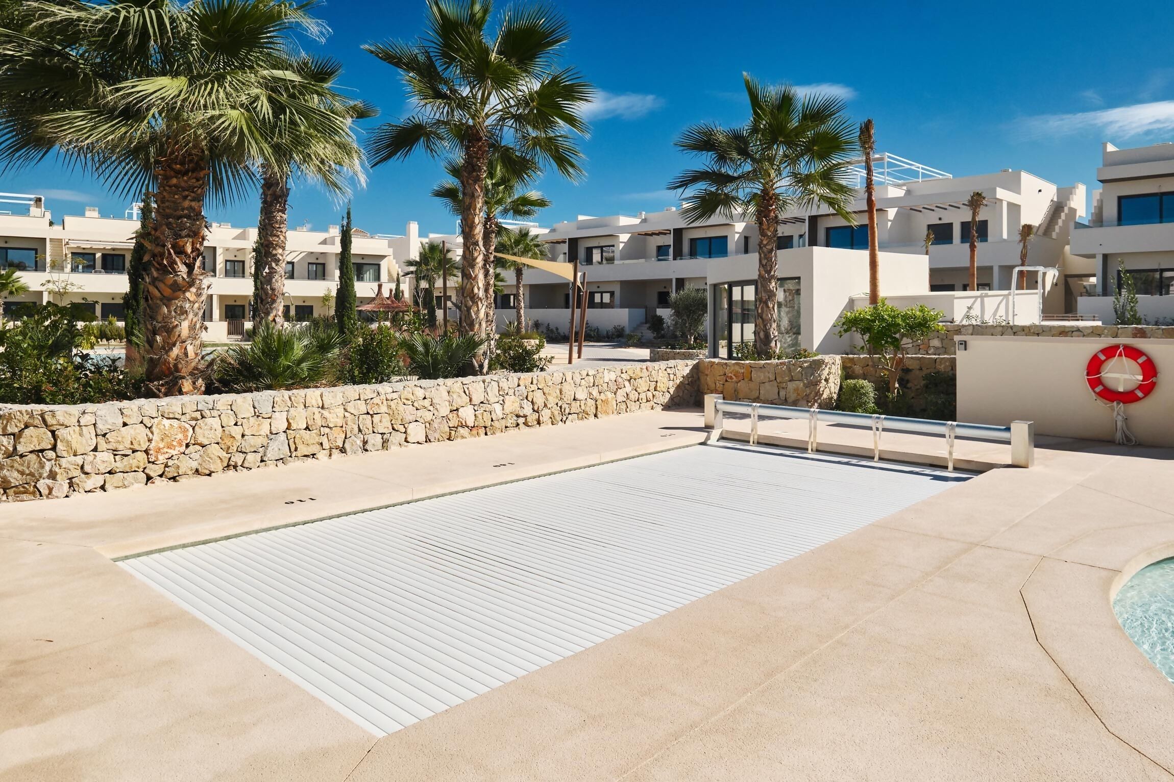 Une zone de piscine équipée d’une bouée de sauvetage, bordée de palmiers, avec des bâtiments modernes en arrière-plan.