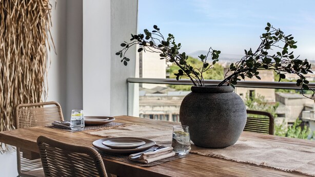 A dining table with a vase of greenery, plates, and glasses, set on a balcony with a view of buildings. - Sanders Old Port Thessaloniki (Thessaloniki)