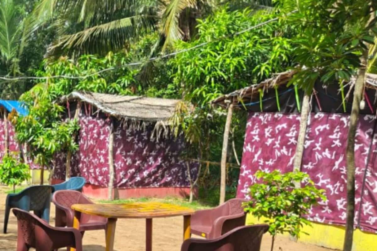 A sandy outdoor area with a table and chairs, surrounded by a thatched-roof structure and a wall with a white pattern.