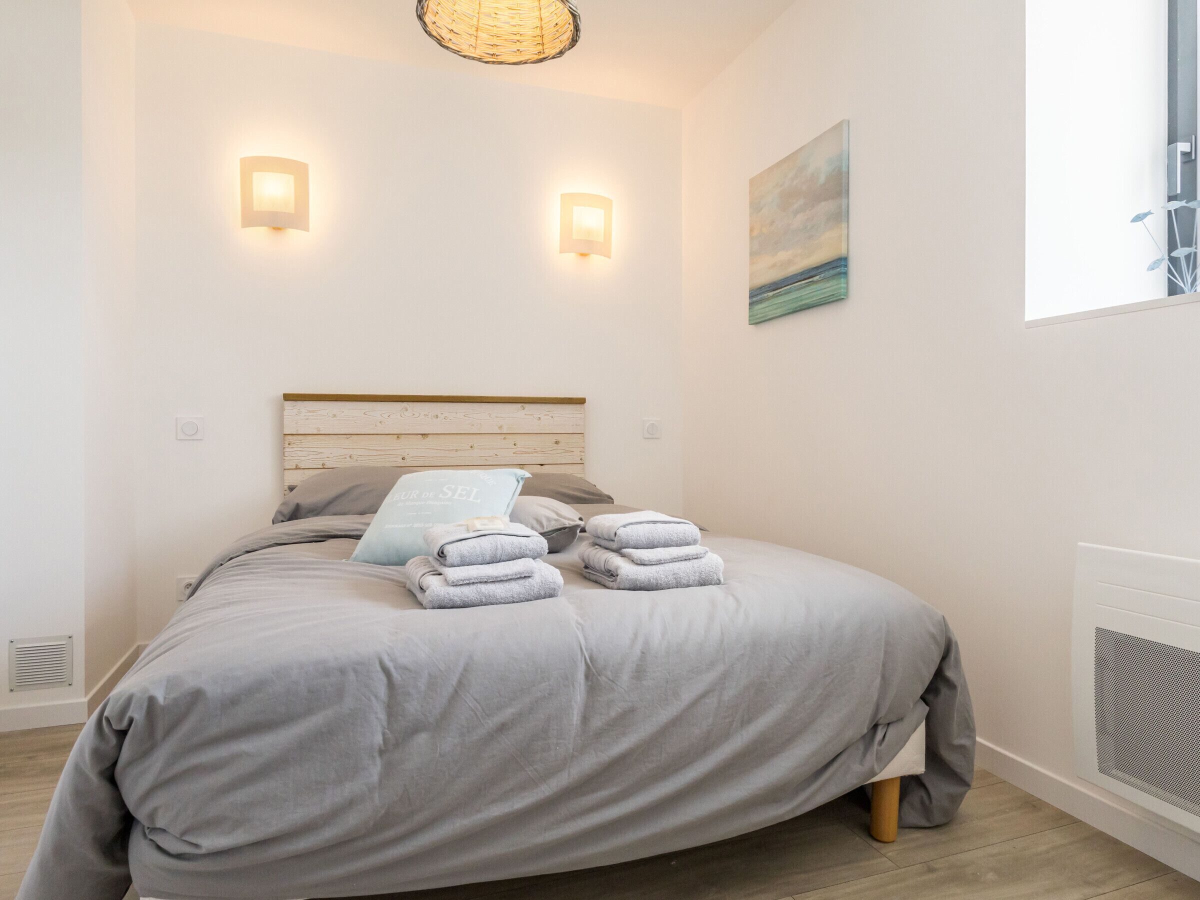 Un lit bien fait, recouvert d’une couvre-lit grise et agrémenté d’oreillers, deux lampes fixées au mur, un tableau et une fenêtre avec des stores.