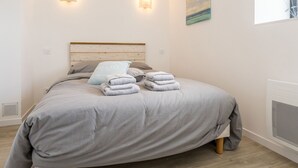 Un lit bien fait, recouvert d’une couvre-lit grise et agrémenté d’oreillers, deux lampes fixées au mur, un tableau et une fenêtre avec des stores.