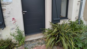 Une porte noire avec une fenêtre au-dessus, une enseigne sur le mur et quelques plantes autour de l’entrée.