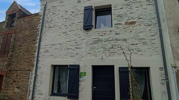 Un bâtiment à deux étages, dont les façades sont en pierre et en crépi, avec deux fenêtres dotées de volets, une porte et un petit jardin agrémenté de plantes.