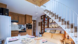Une cuisine chaleureuse avec des meubles en bois, une table à manger dressée avec des assiettes, des verres et un pichet, et un escalier en colimaçon menant à une chambre en mezzanine équipée d’un canapé.