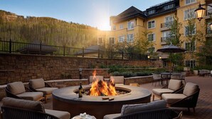 A circular outdoor seating area with a fire pit, surrounded by wicker chairs and tables, set against a backdrop of a multi-story building and a forested hillside. - RITZ CARLTON RESIDENCE - Vaill Colorado,  3 bedroom, lovely mountain views . (Vail)