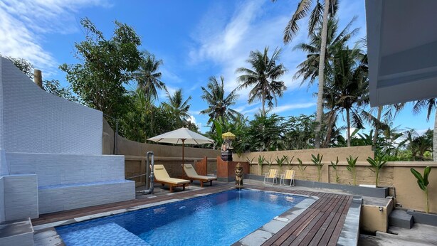 A swimming pool with wooden deck and lounge chairs, surrounded by palm trees and a clear sky. - Rai Wood Carver and Homestay (Ubud)