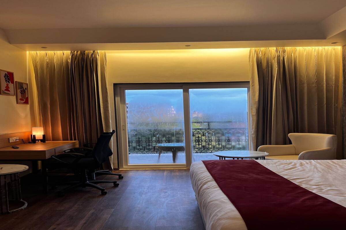 A hotel room with a bed, a desk, a chair, a small table, and a view of a cityscape through a large window.