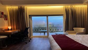 Habitación de hotel con cama, escritorio, silla, mesita y vista a un paisaje urbano a través de un ventanal grande.