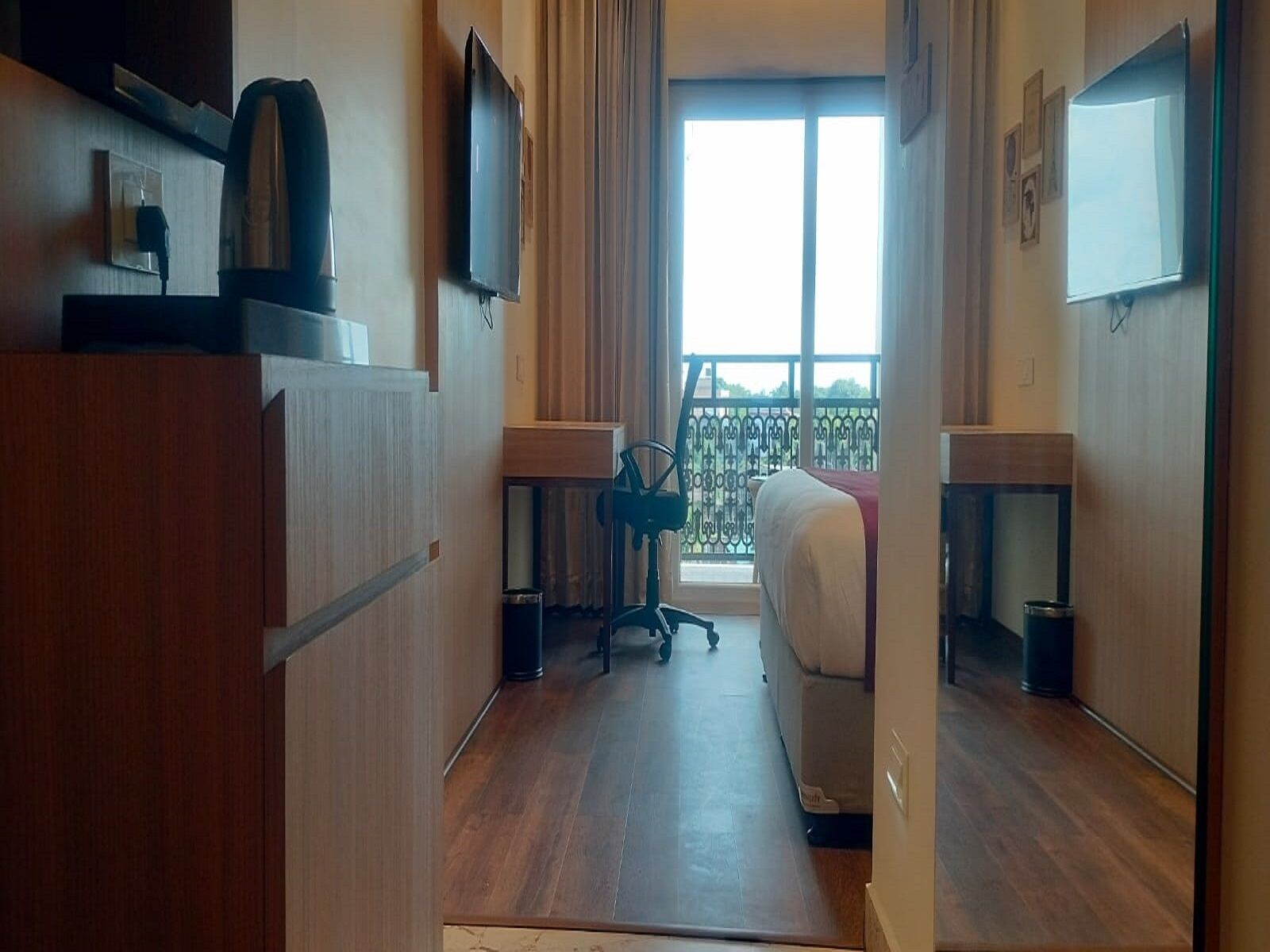 A hotel room with a bed, a desk, a chair, a TV, and a balcony with a railing.