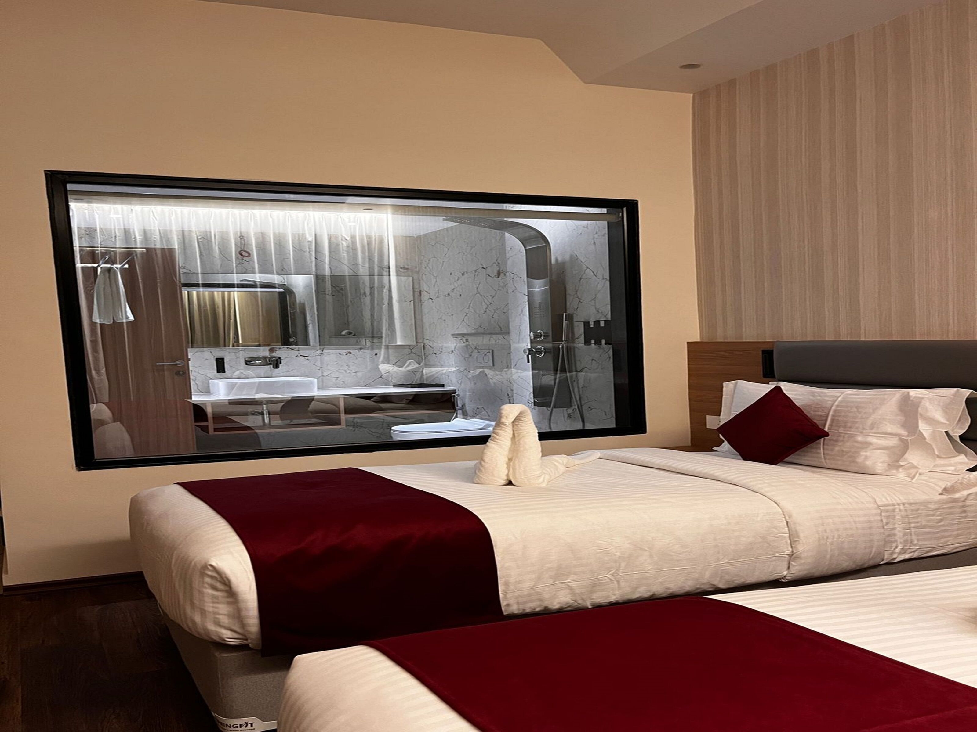 A hotel room with a large window, a bed with white and red bedding, a wooden headboard, and a marble bathroom visible through the window.