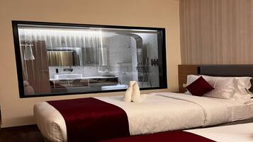 A hotel room with a large window, a bed with white and red bedding, a wooden headboard, and a marble bathroom visible through the window.