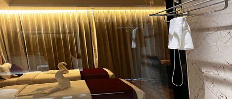 A hotel room with a large window, a bed with a red accent, a towel rack, and a marble wall.
