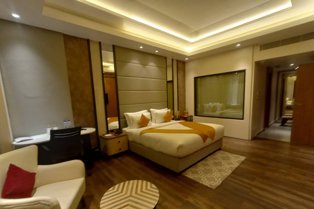 A modern hotel room with a large bed, a sitting area with a chair and a small table, a bedside table with a lamp, and a large window.