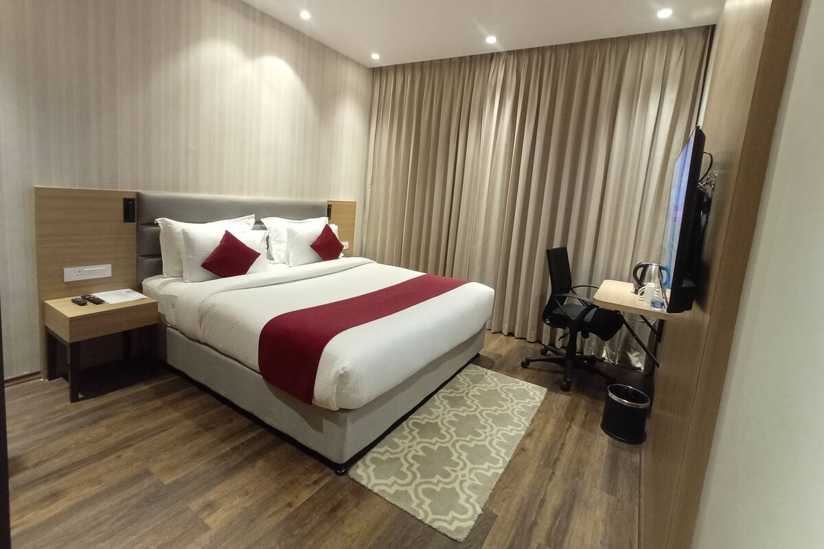 A modern hotel room with a large bed, a desk with a chair, a television, and a small table with a lamp.