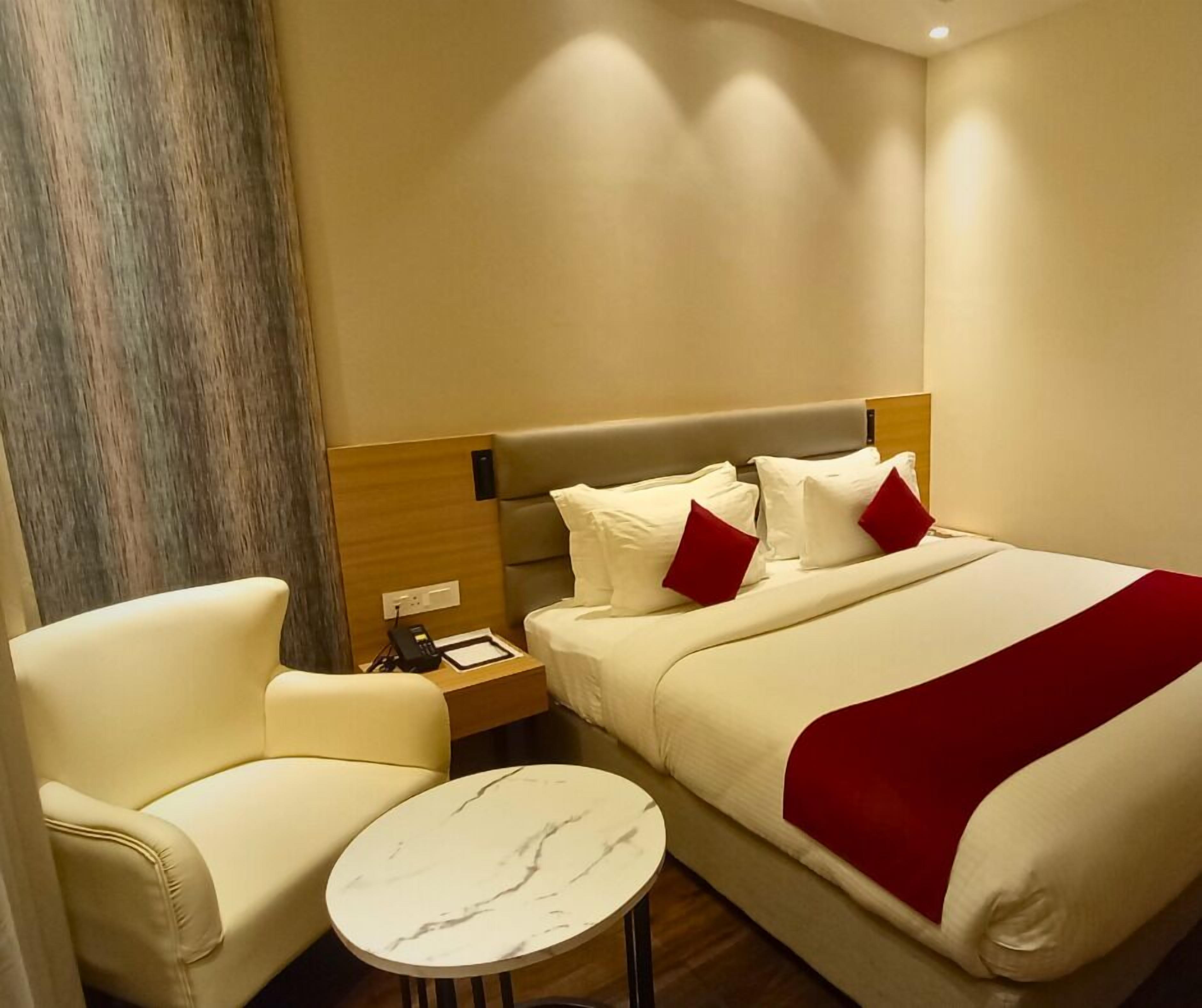 A hotel room with a bed, a chair, a small table, and a nightstand with a phone and a lamp.