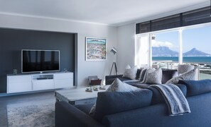 A modern living room with a large flat-screen TV, a sofa set, a coffee table, and a floor lamp. There is a framed picture on the wall and a view of the sea and mountains through the window.