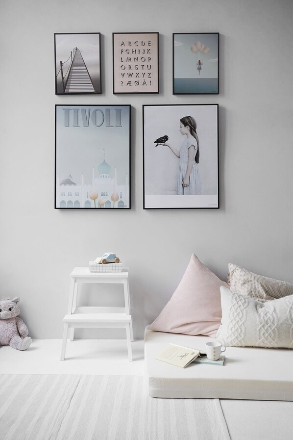 A minimalist room with a white sofa, a small white table, and a teddy bear. There are three framed pictures on the wall, including one with a mosque and another with a girl holding a bird.
